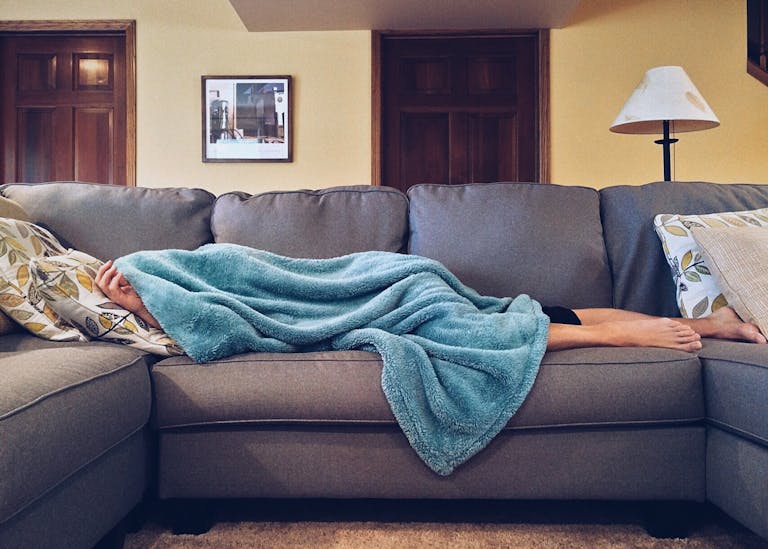 Person napping under a blue blanket on a comfortable couch in a modern living room setting.